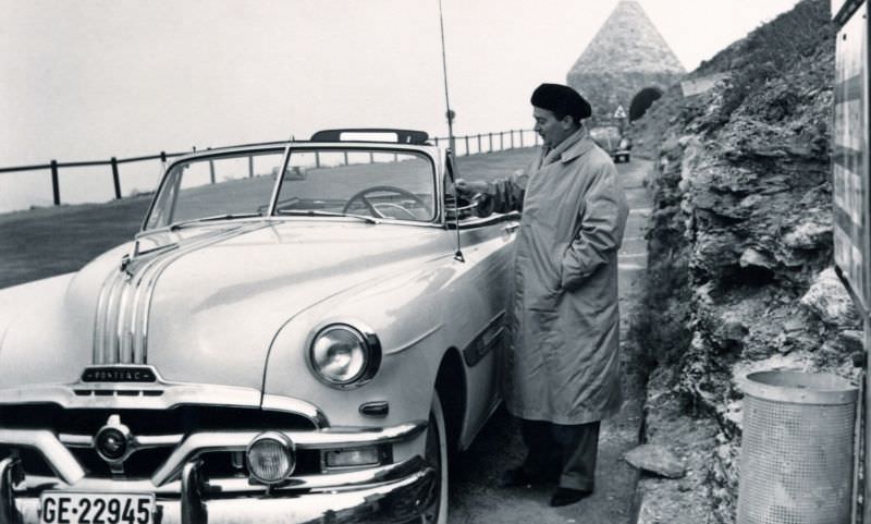 A fellow wearing a Basque-style beret and a raincoat posing with a 1952 Pontiac Chieftain Convertible on an Alpine road.