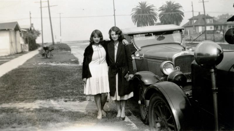 Two young ladies, possibly sisters, posing with a 1928 Pontiac in a suburban street.