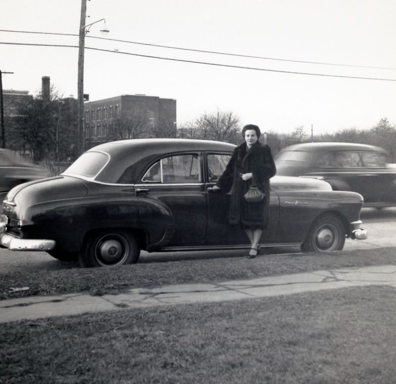 A middle-aged lady in a fur coat posing at the passenger side door of a 1950 Pontiac Chieftain Sedan on a bleak winter's day, 1952
