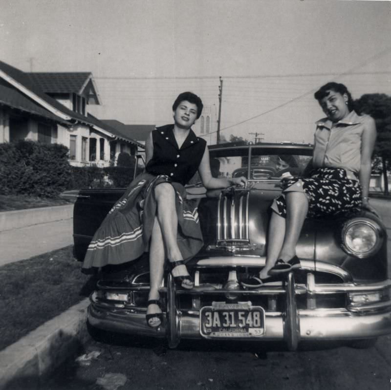 Two young Latinas posing on the hood of a 1950 Pontiac Chieftain Convertible.
