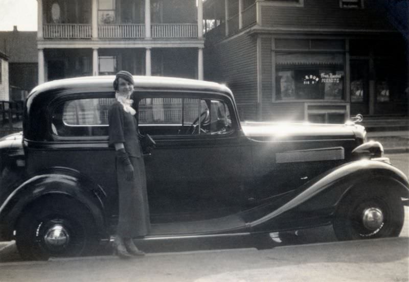 An elegant lady wearing a dark female suit, opera gloves and a white jabot-style collar posing with a shiny 1934 Pontiac Economy Eight Sedan in front of a row of timber houses, 1934