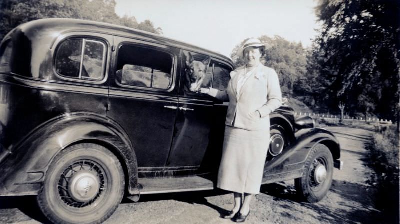 A middle-aged lady and her German shepherd posing with a 1934 Pontiac Economy Eight Sedan in the countryside.