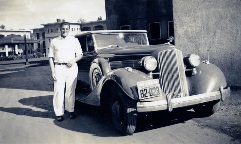 A dapper fellow dressed in a white shirt and white slacks posing with a 1934 Pontiac Convertible in an exotic location.