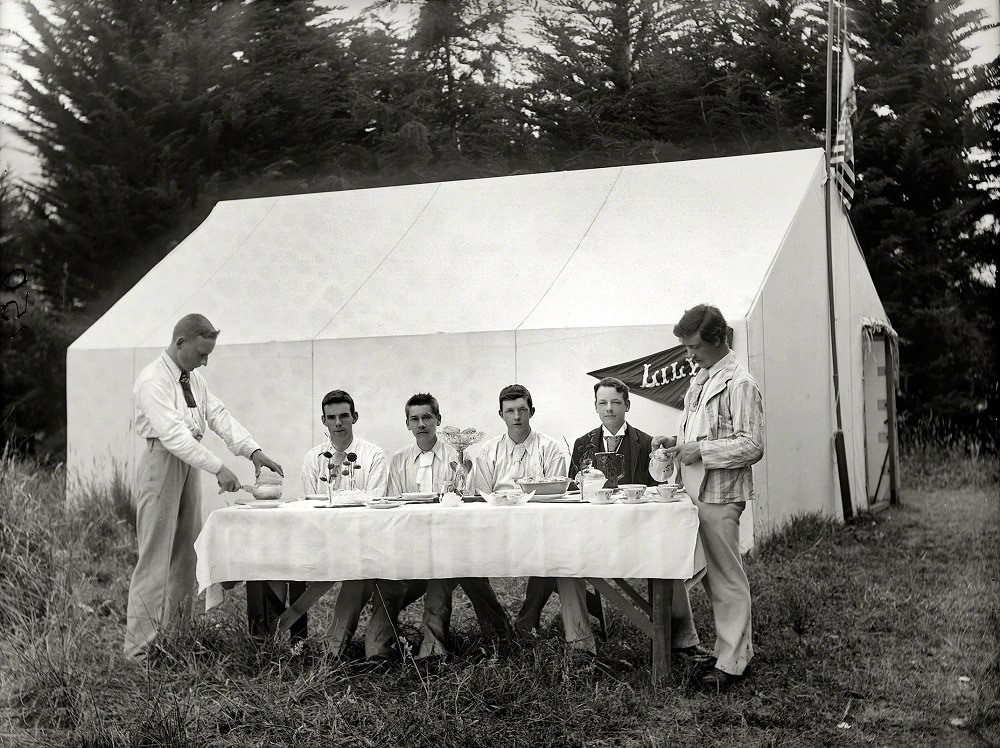 Young men in formal wear at table ready to have a meal, next to tent 'Lily' at camp site, probably in Sumner, Christchurch, New Zealand, 1910