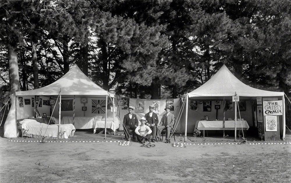 Unidentified men sitting between two open tents with beds, tables, pictures, flags and posters on the walls, New Zealand, 1910
