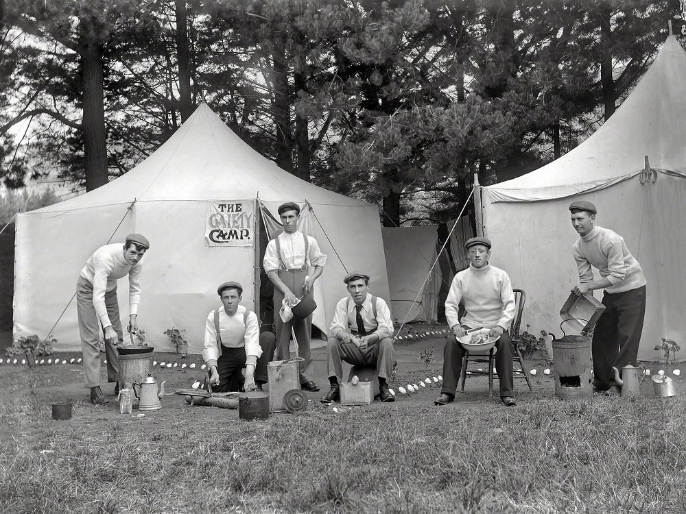 Group men outside a tent with a sign reading 'The Gaiety Camp,' showing each man performing domestic duties, Christchurch district, New Zealand circa 1910