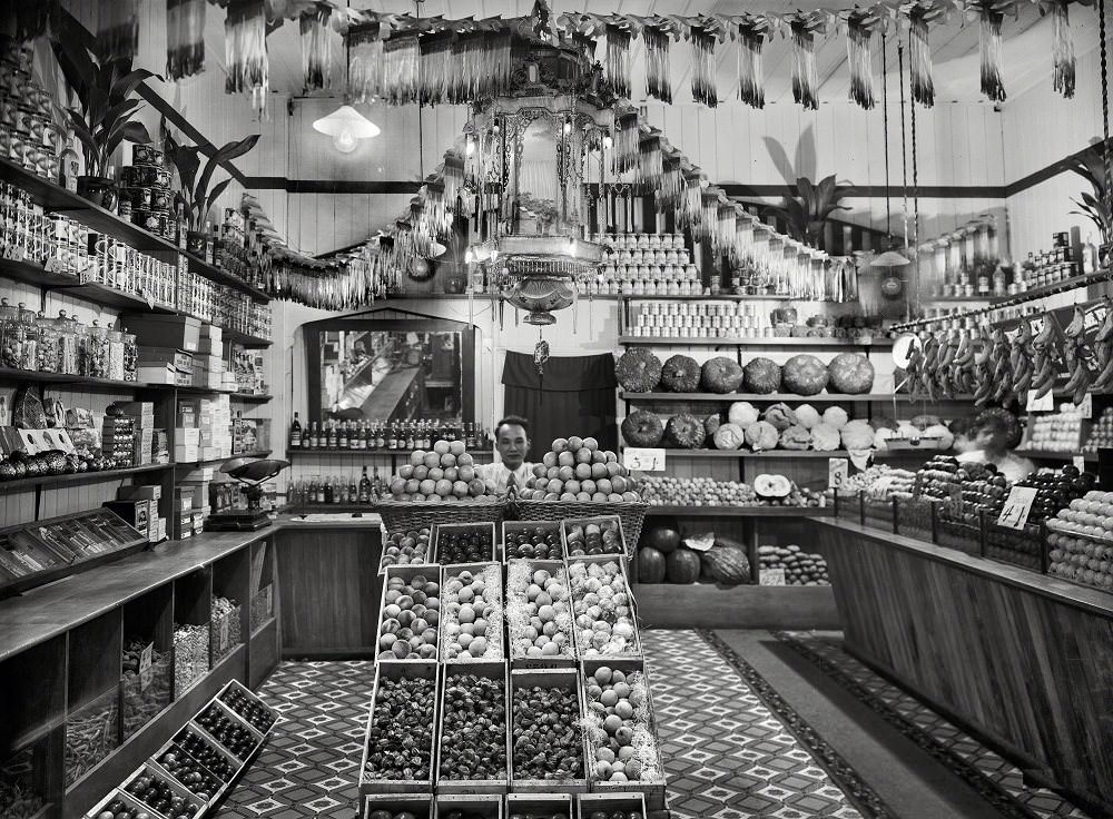 Greengrocery, Chinese shopkeeper with baskets of apples and boxes of peaches, cape gooseberries and other fruits, Taranaki region, New Zealand, 1920s