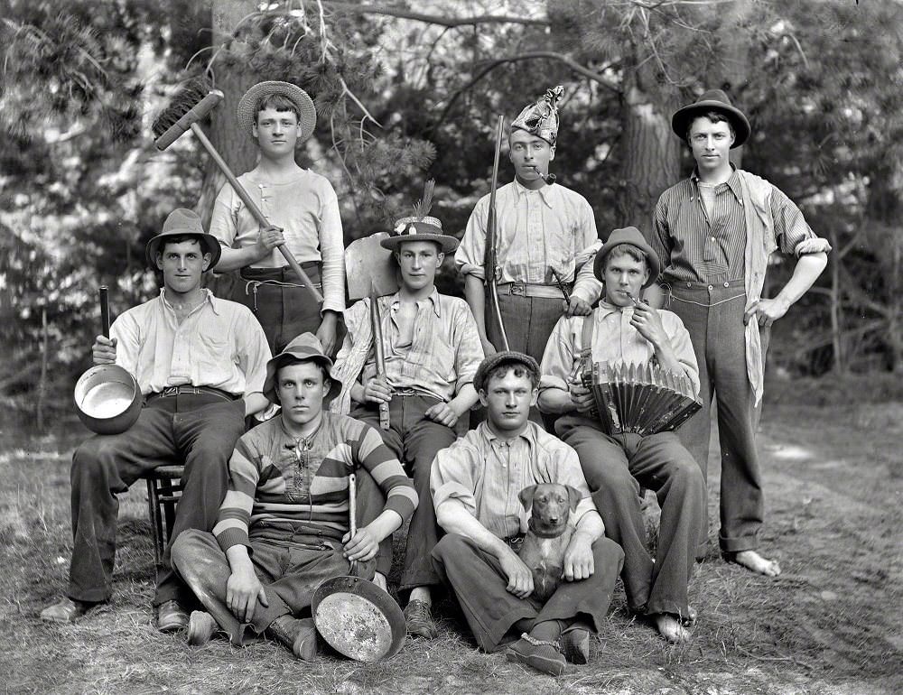 Eight young men in ragtag clothing with hats, pots, broom, shovel, accordion, pipes, gun and dog, Christchurch, New Zealand circa 1905