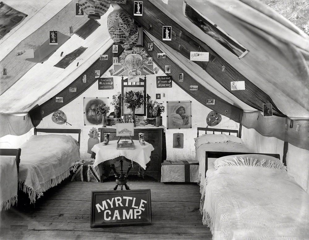 Interior of large tent decorated with posters and picture postcards, with tallboy and mirror, trunks with flags, lamps and 'Myrtle Camp' sign, Christchurch, New Zealand circa 1905