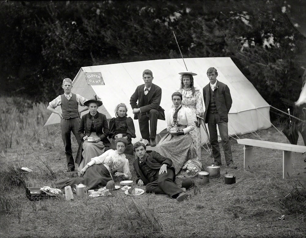 Young people with camping gear, having tea and cake in front of tent with 'Clover Camp', near Christchurch, New Zealand, 1905