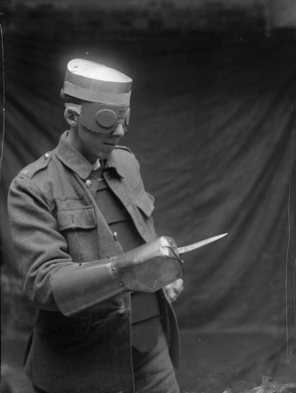 A man demonstrates a steel cap, splinter goggles (vision is obtained through thin slits in goggles), and a steel dagger gauntlet, manufactured for the British military during World War I.