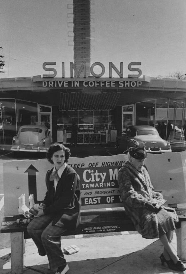 Two women sit on a bench outside a Hollywood drive-in restaurant.