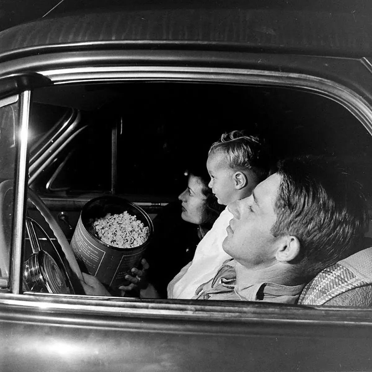 A late night at the drive-in, a couple and their young son watch a film while they sit in their ’41 Buick coupe, with almost an equally large canister of popcorn.