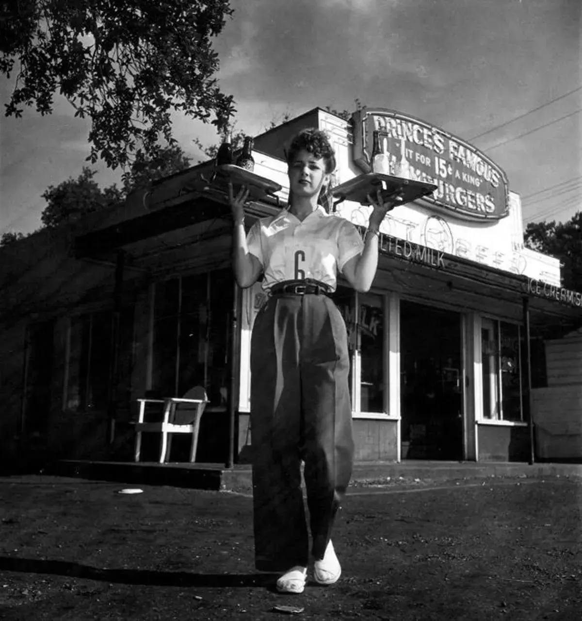A carhop holds two trays as she poses for a shot in front of a small diner, Houston, Texas, 1945.