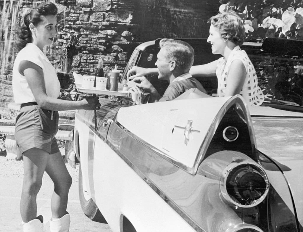 A waitress serves food to a passenger and driver of a classic car complete with tail fins.