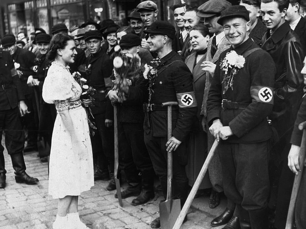 Members of the German Labor Corps stand along a street curb holding shovels as a young girl welcomes them to Sudetenland with flowers.