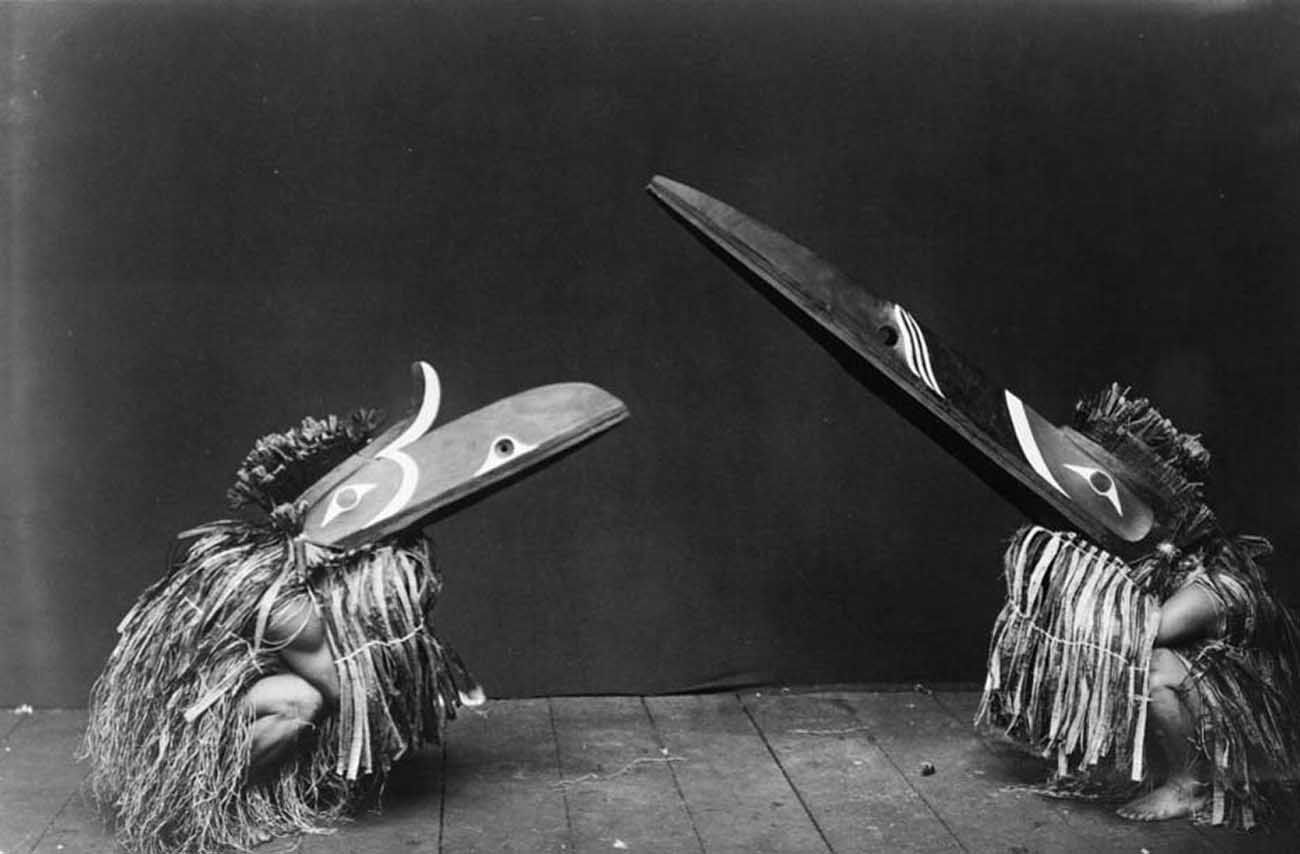 Nakoaktok men in ceremonial dress, with long beaks, crouching on their haunches.