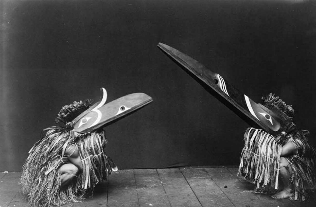 Nakoaktok men in ceremonial dress, with long beaks, crouching on their haunches.