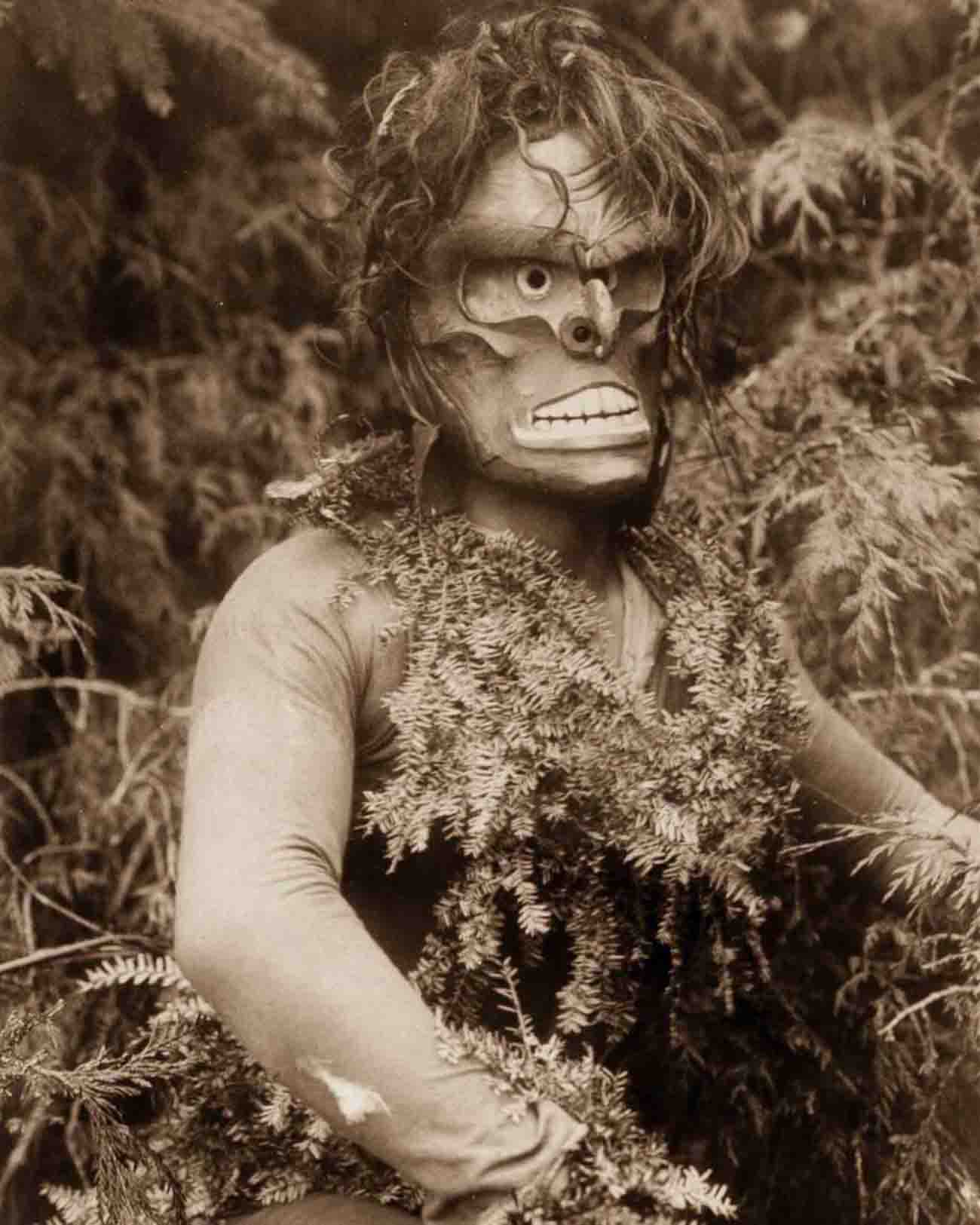 Dancer representing Paqusilahl (“man of the ground embodiment”), wearing a mask and shirt covered with hemlock boughs, representing paqus, a wild man of the woods.
