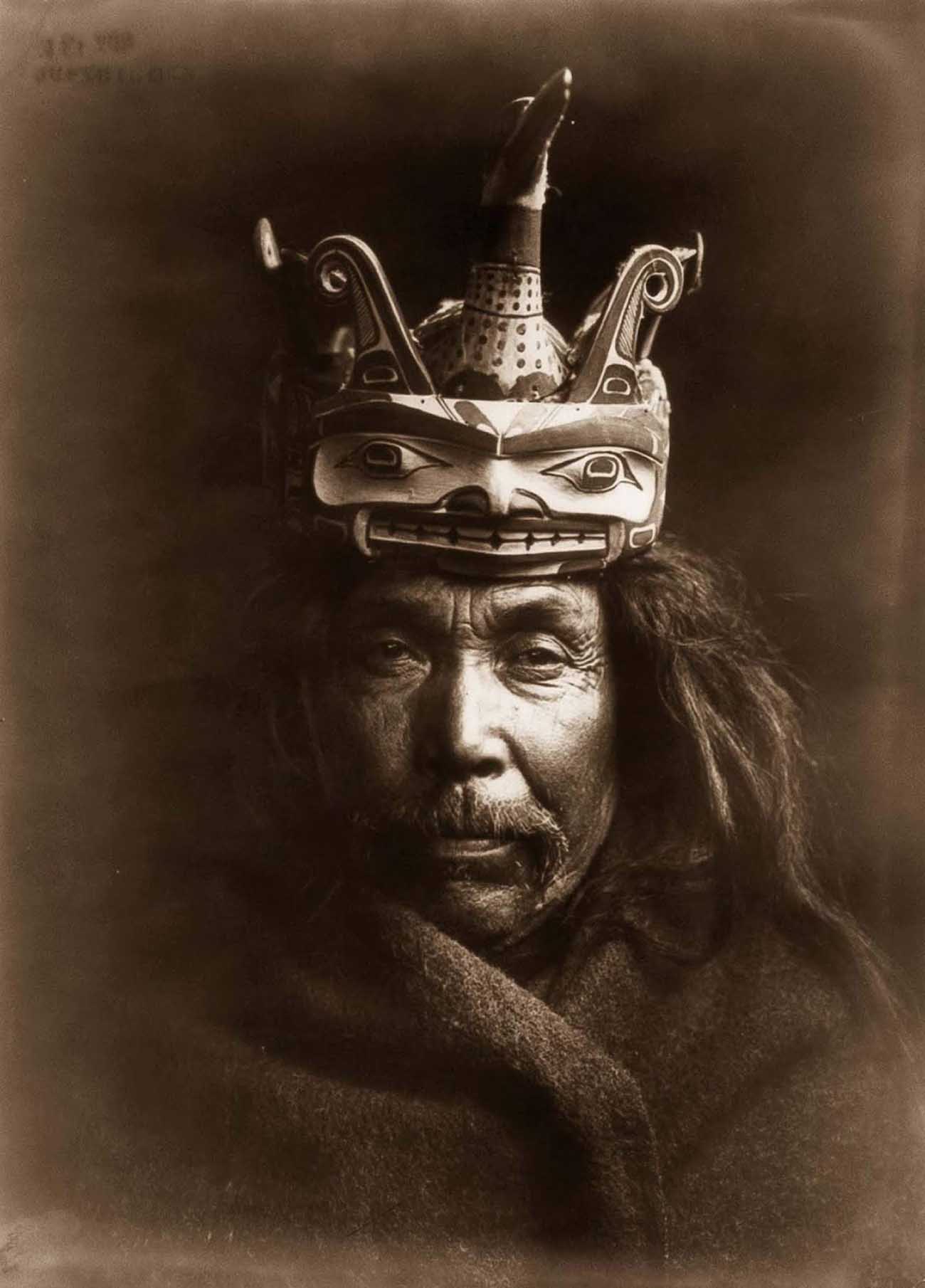 Kwakiutl man, head-and-shoulders portrait, facing front, wearing a mask depicting a loon on top of a man’s head to facilitate the loon changing into the form of a man.