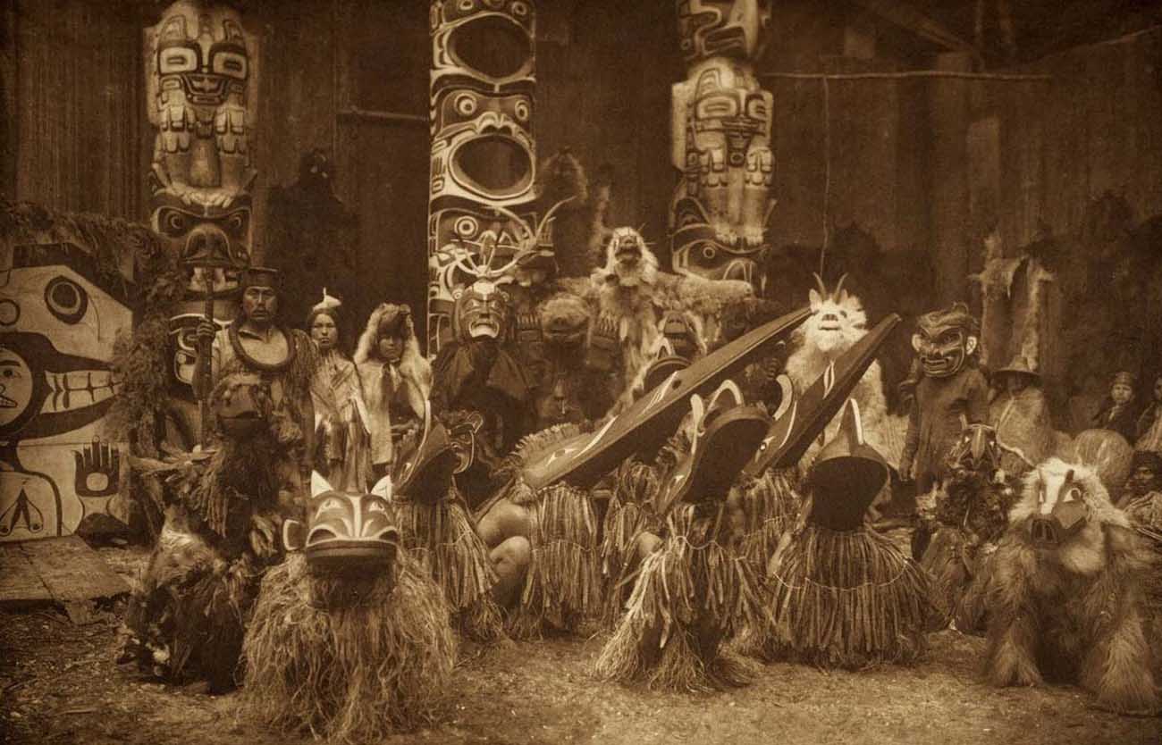 During the winter ceremony, Kwakiutl dancers wearing masks and costumes, crouch in foreground with others behind them. The chief on the far left holds a speaker’s staff.