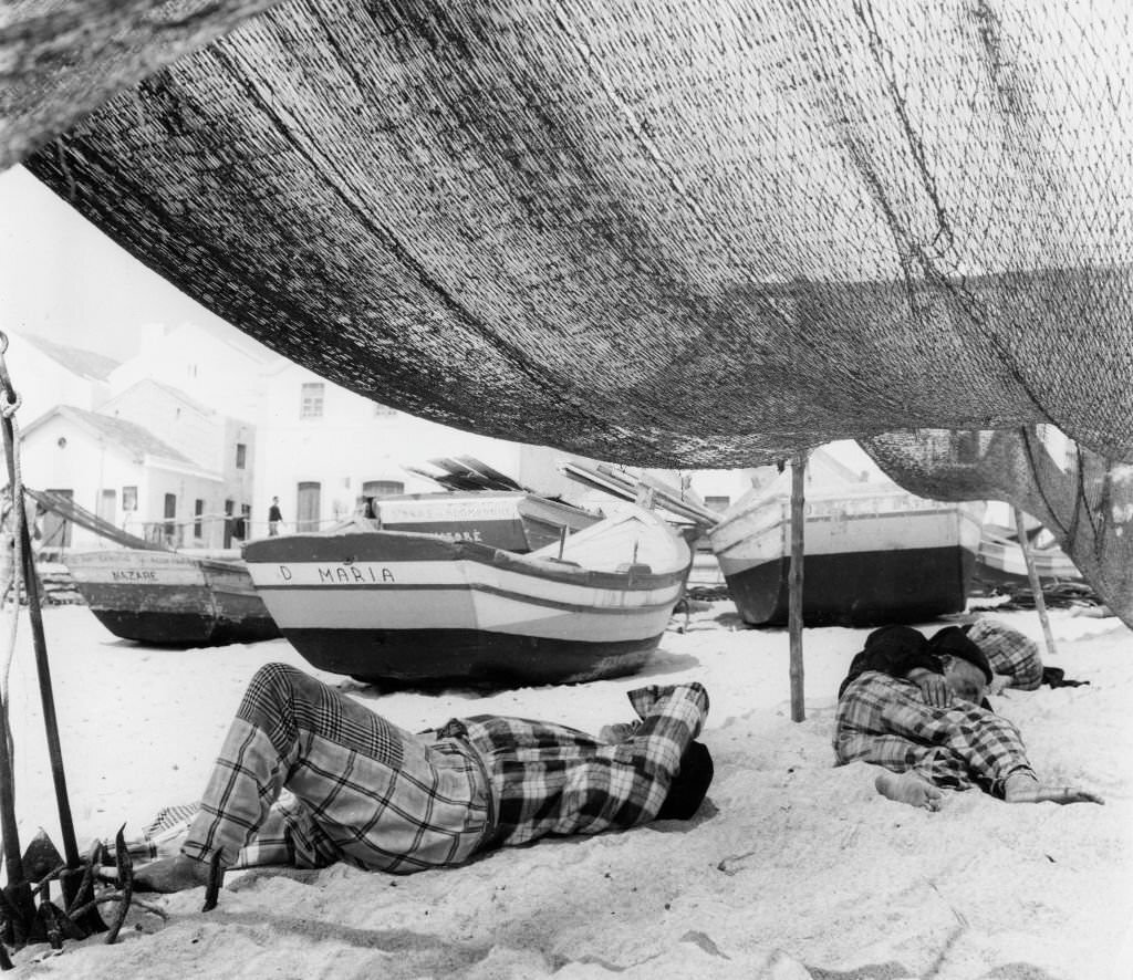 Portugeuse fishermen in traditional checks having a siesta underneath their nets on the beach at Nazare, 80 miles north of Lisbon, 1958