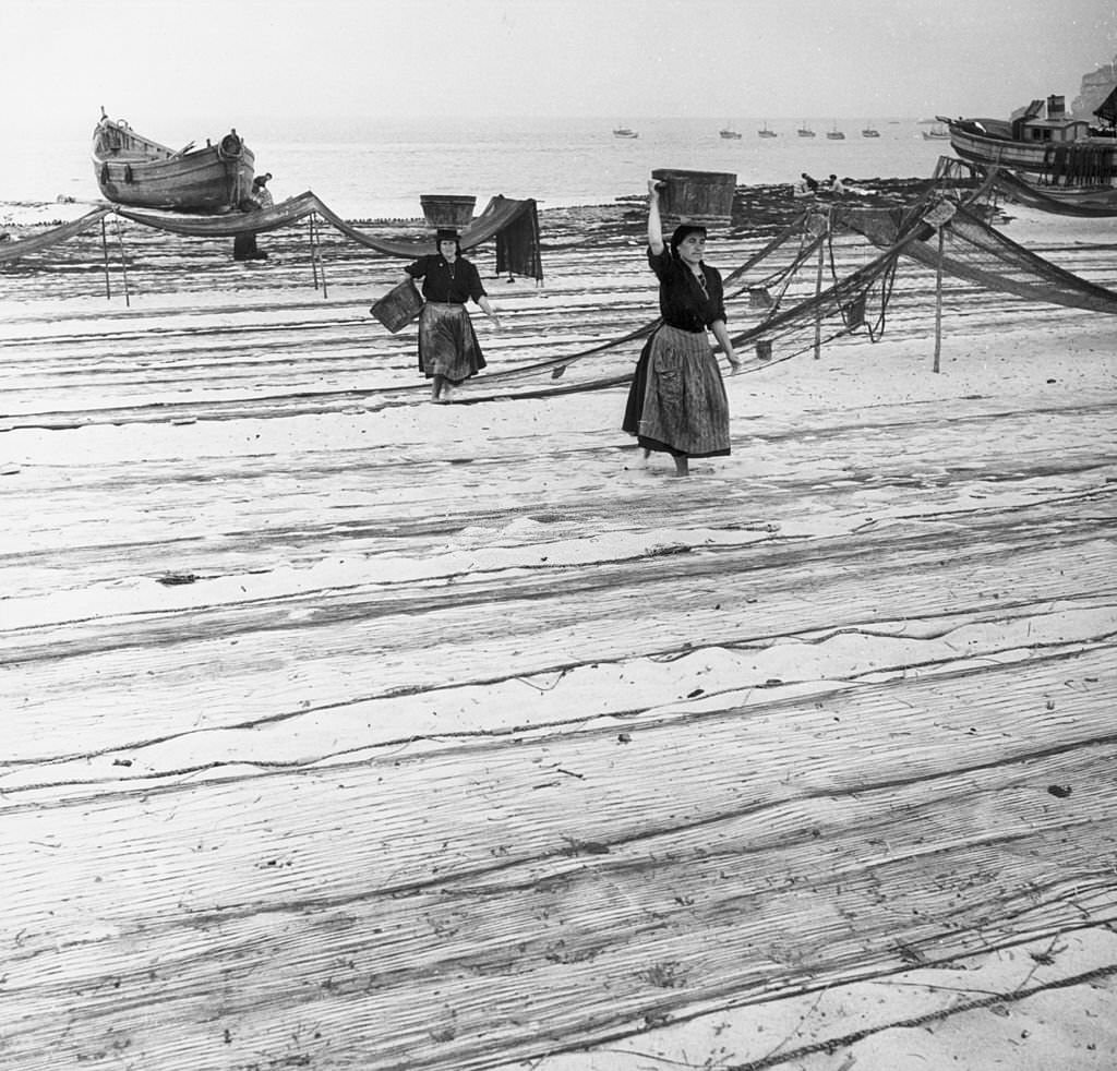 Women carrying fish walk across the drying fishing nets on the beach at Nazare.