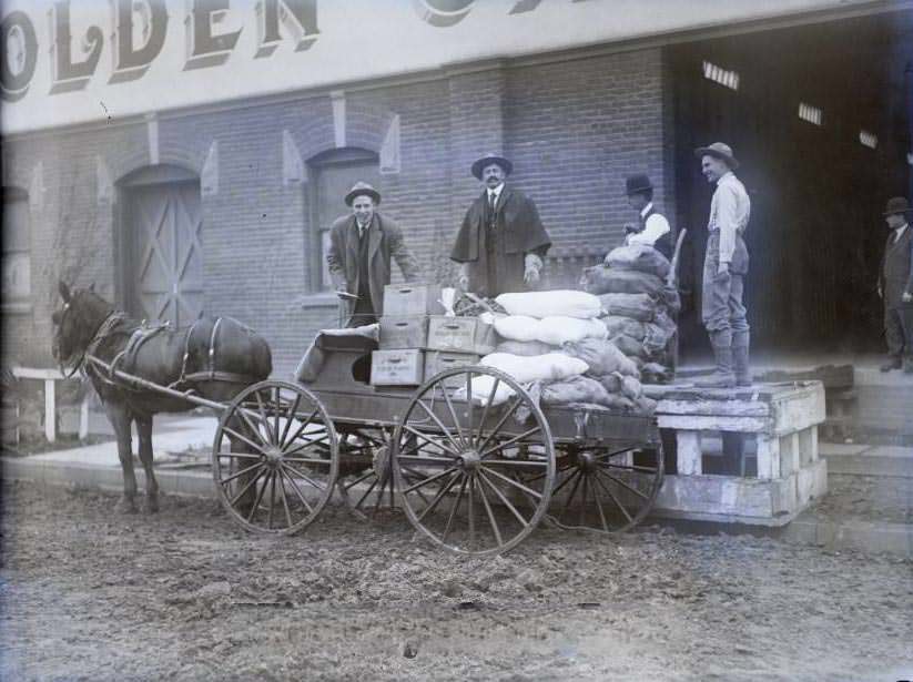 Horse-drawn wagon being loaded in front of brick building, 1870s