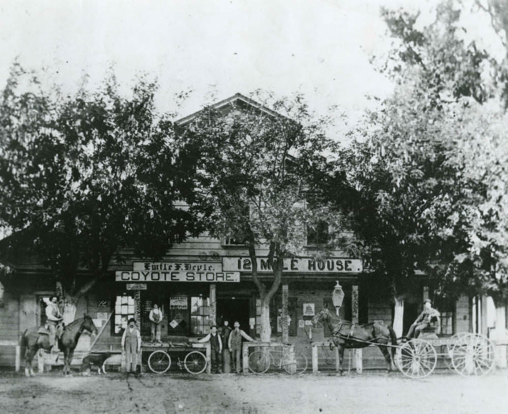 Several men pose with their bicycles and horses in front of Twelve Mile House and Coyote Store, 1890