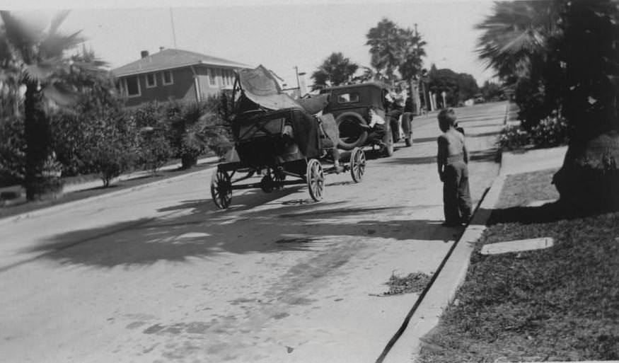 Vehicle towing older car as young boy looks on from the curb, 1932