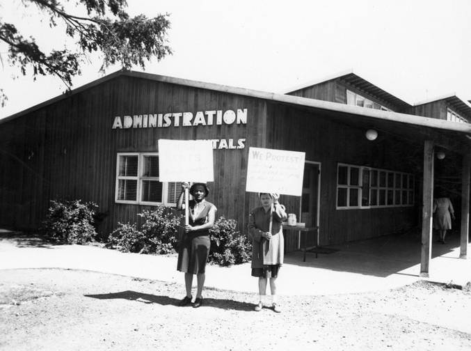 Two women, one African-American and one Caucasian, hold protest signs outside a Vancouver Housing Authority building, 1940s