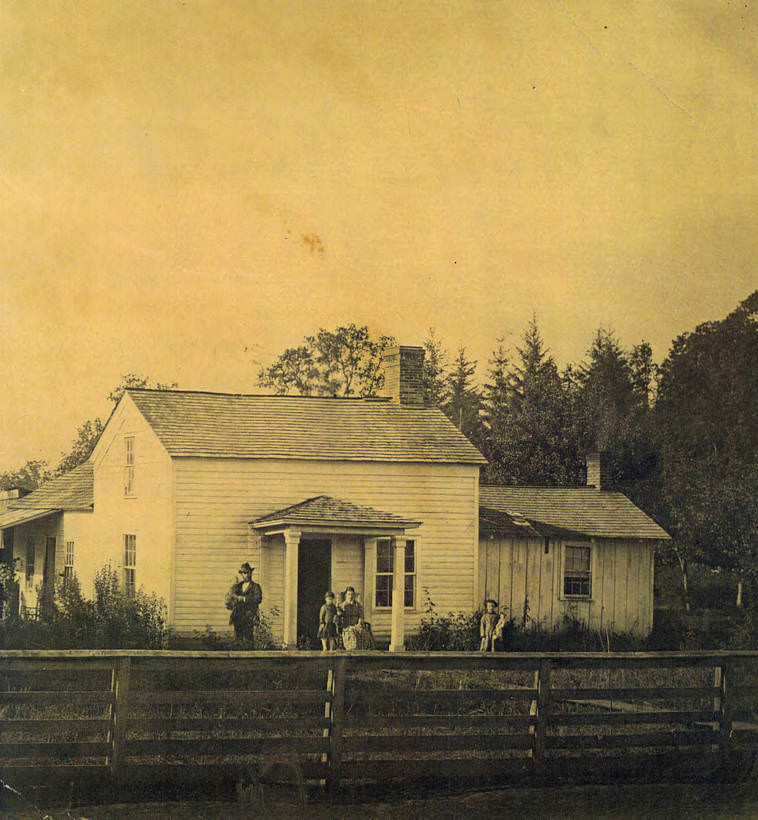 Wooden home with fenced yard. A man holding an infant stand to the left of the porch, 1883