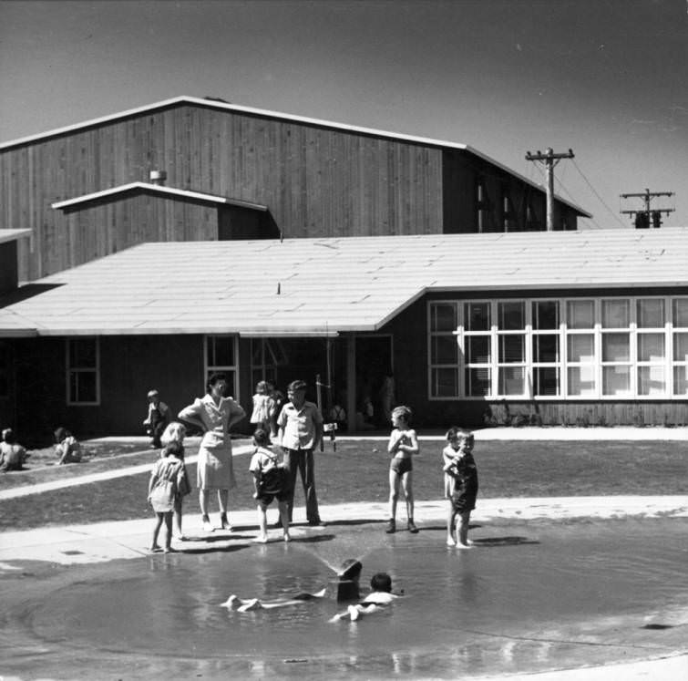 Children play in a wading pool outside a building. V.H.A, 1950s