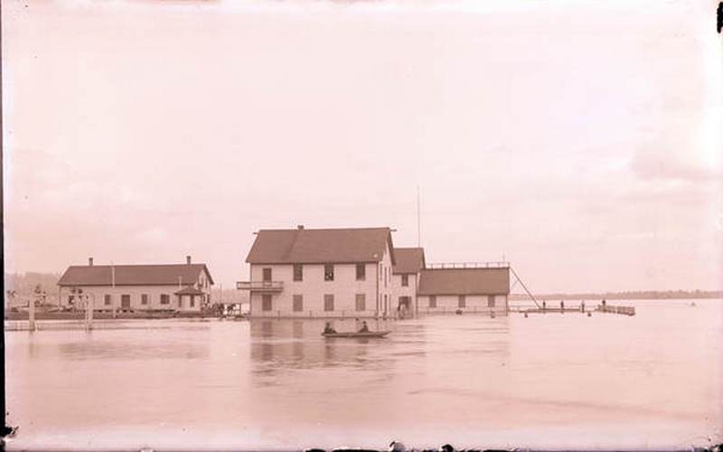 Two people in a boat travel through a flooded area of Vancouver, 1893
