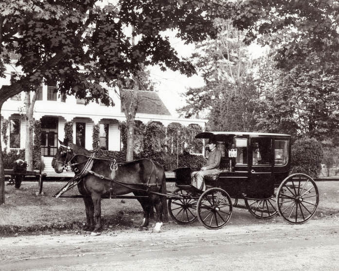 A black carriage with a team of horses and a driver in an overcoat and top hat sits in front of a house on Officers Row, 1890s