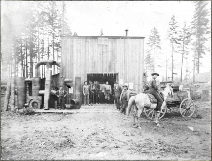 Men stand before a blacksmiths shop with pipes and conduits off to the side, 1904