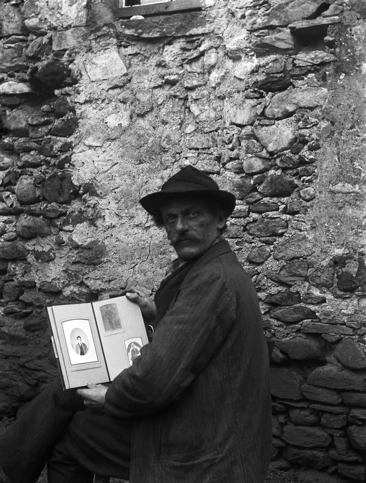 Self-portrait of Roberto Donetta with hat and a photo album in hand, in front of a wall, Bleniotal