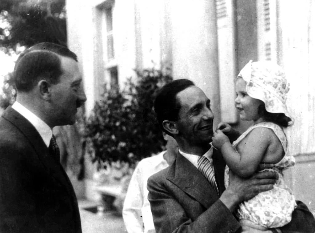 German Chancellor Adolf Hitler, left, chats to his Propaganda Minister Dr, Goebbels who is holding his daughter Helga, during Hitler+s visit to their home in Berlin, 1934