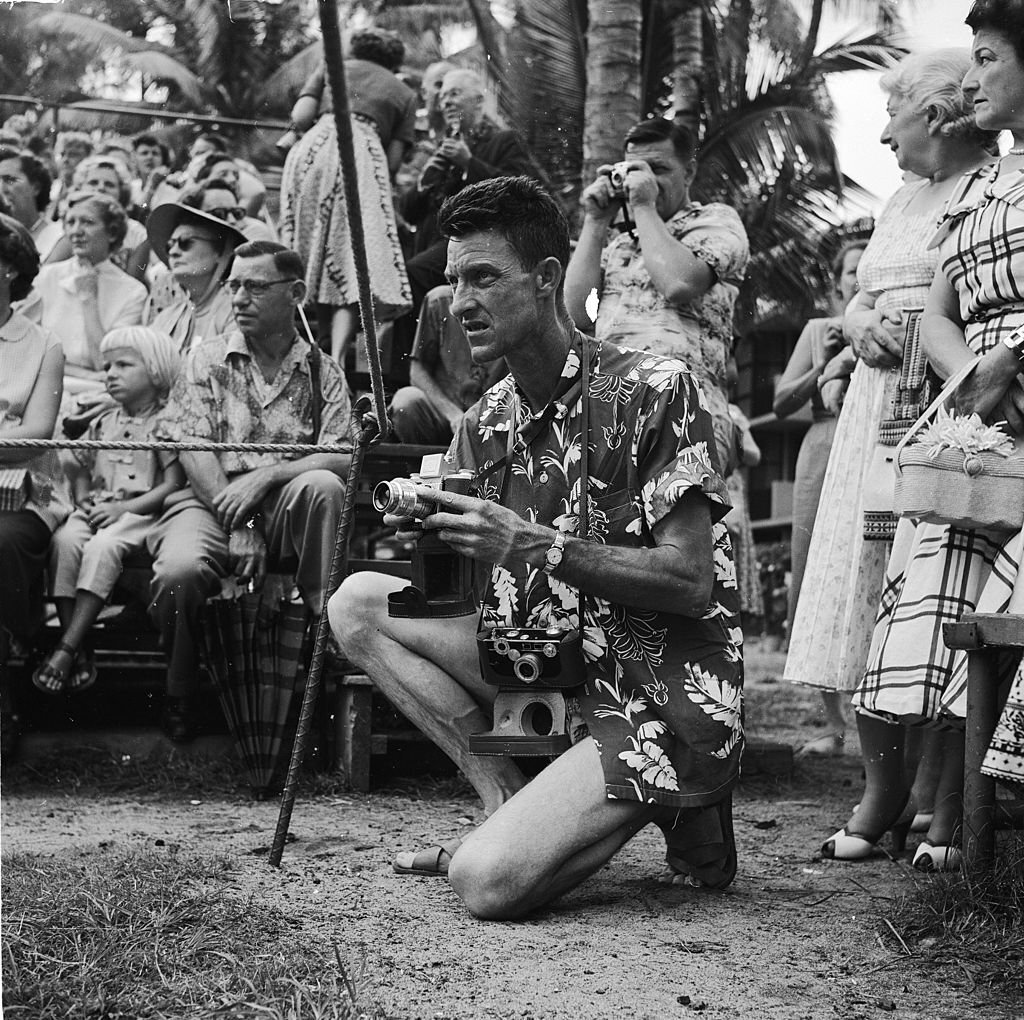 A tourist wearing the traditional Hawaiian shirt kneels with his camera at the ready, awaiting the arrival of the Hula dancers on Honolulu's Waikiki Beach, in Hawaii, 1953