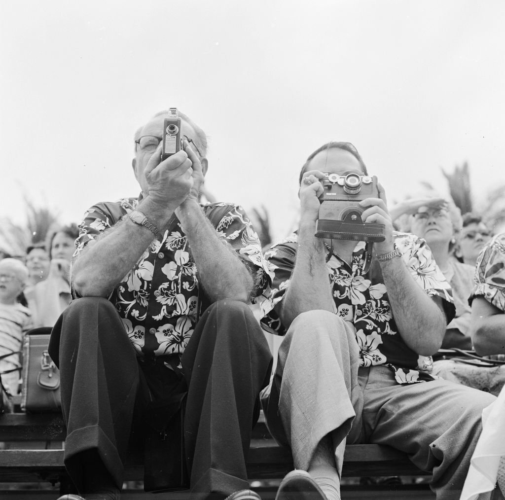 Two tourists in matching Hawaiian shirts poise with their cameras at the ready, awaiting the arrival of the Hula dancers on Honolulu's Waikiki Beach, in Hawaii, 1953