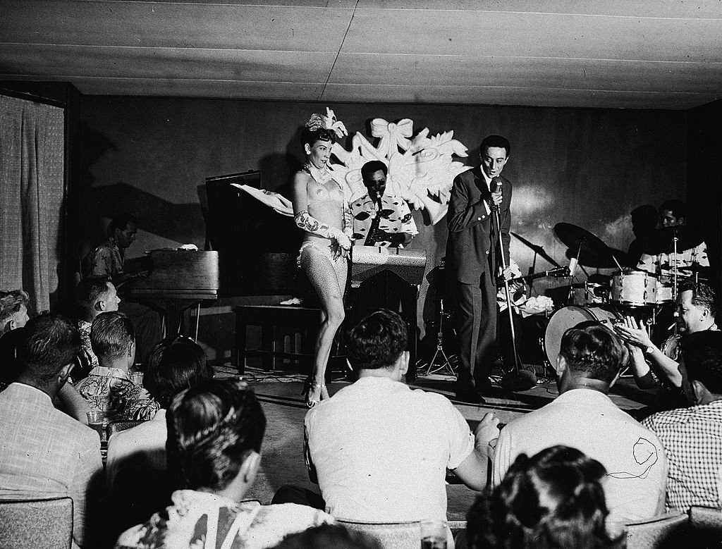 American comedian Lenny Bruce performs on stage with exotic dancer Windee Gayle and a saxophonist at the Orchid Room, Waikiki, 1950s.