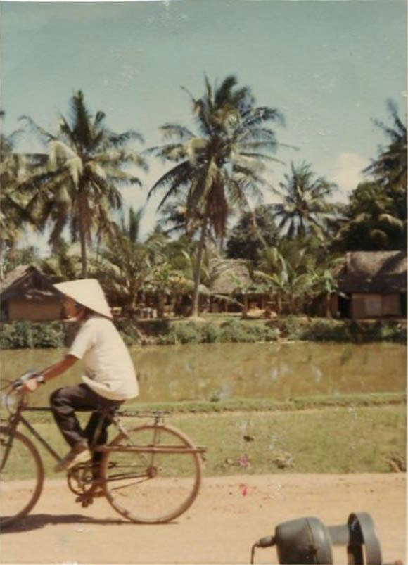 An unidentified Vietnamese village and coconut trees, with a young Vietnamese boy riding past on a bicycle somewhere in South Vietnam.
