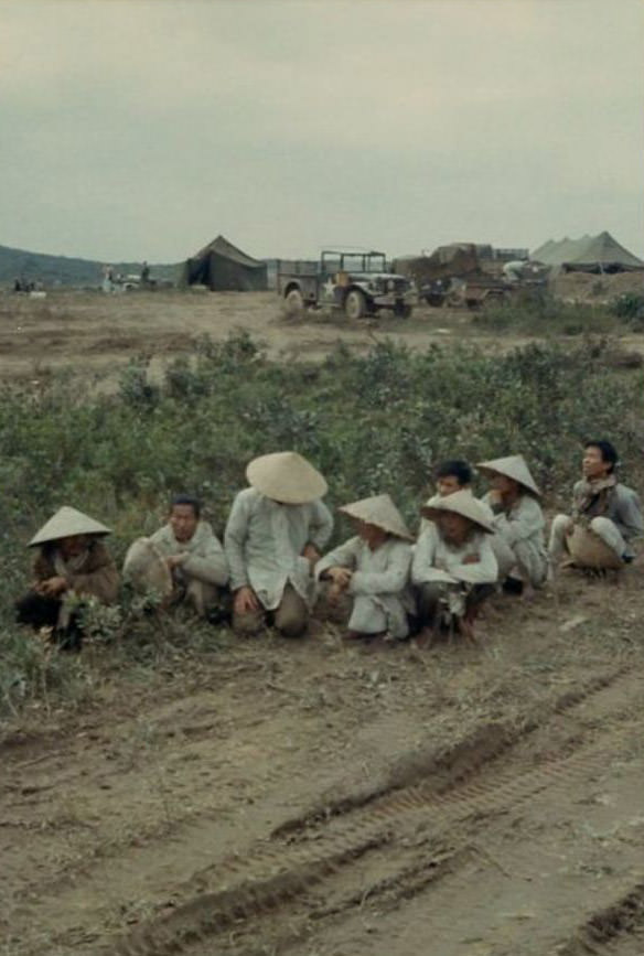 Eight Vietnamese people wearing conical hats, kneeling or sitting down along a dirt road in the 2nd Battalion.