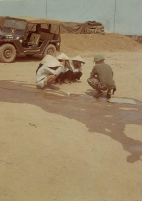 Several Vietnamese people wearing conical hats, kneeling down on a dirt road, as an Army soldier talks to them at the 2nd Battalion.