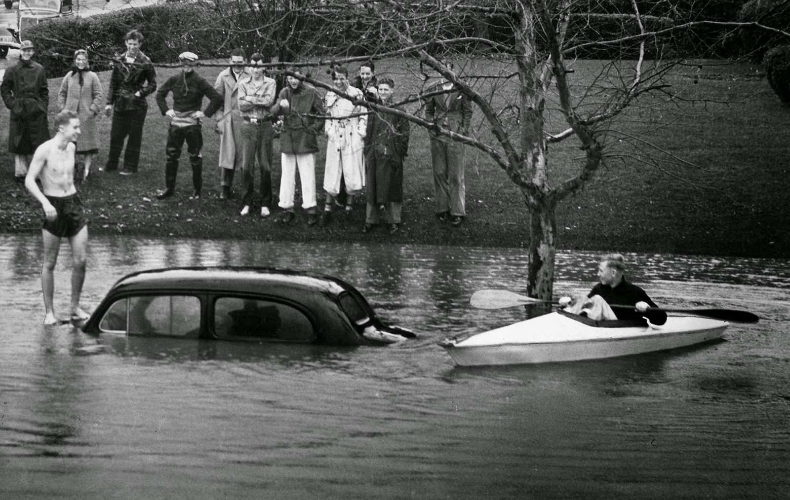 Hugh Beckly stands atop a car almost covered by water at 6th Street near June. Arden Day cruises by in a skiff, 1938