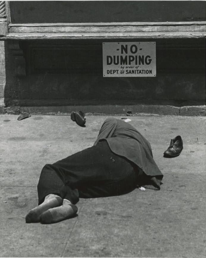 A man sleeps on the ground under a sign that reads NO DUMPING by order of Dept. of Sanitation on the Lower East Side.