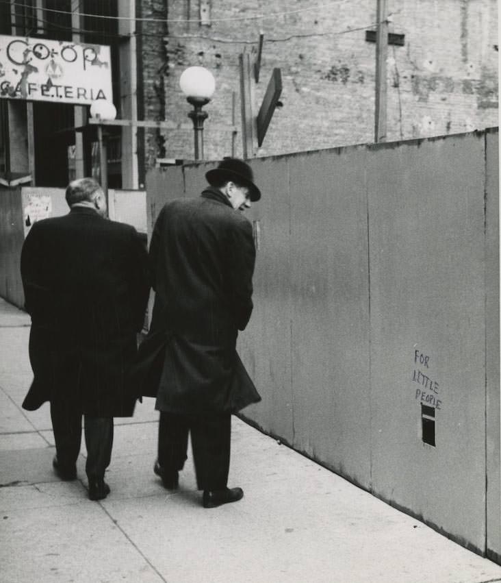 Two men walk by a wooden wall with a rectangular cutout that says FOR LITTLE PEOPLE on the street in New York City. Inscription on verso- For Sidewalk Superintendents.