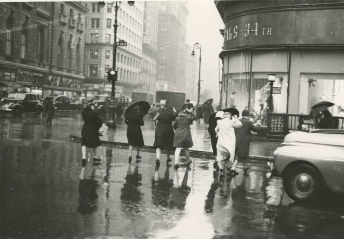 Pedestrians cross the street in the rain towards Saks – 34th store on West 34th Street in Midtown Manhattan