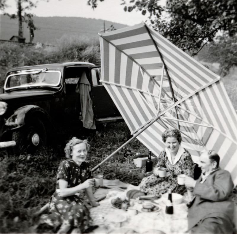Two cheerful ladies and a bald fellow in a suit enjoying a picnic in the countryside, 1955