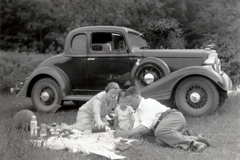A family of three enjoying a picnic in the countryside. A 1933 Pontiac Eight Coupe can be seen in the background, 1935